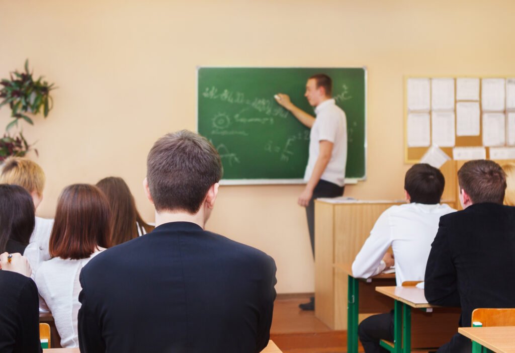 Teacher writing on a chalkboard while students sit and listen in a classroom.